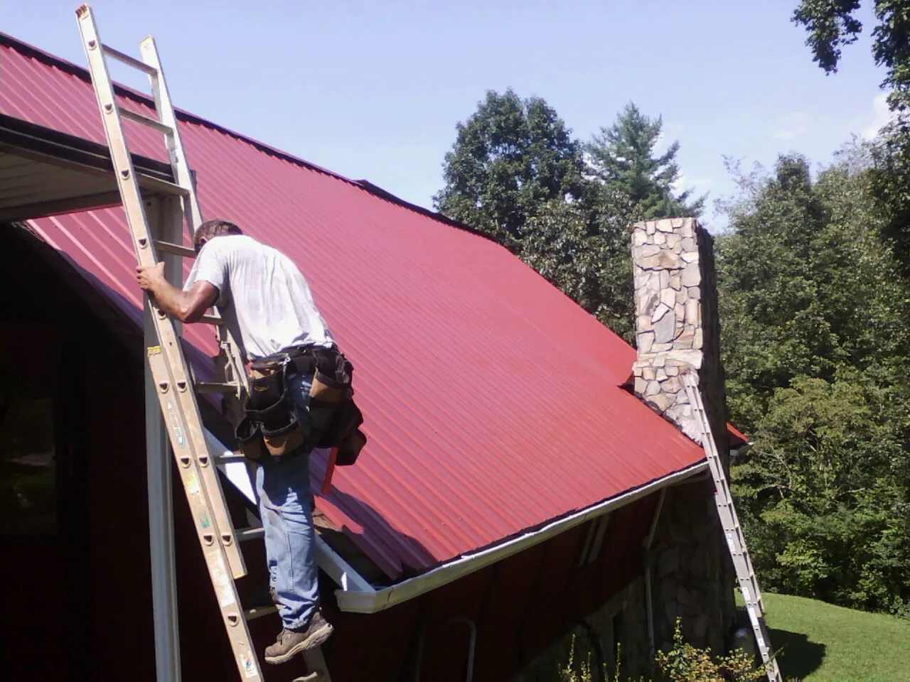 Roofer on a ladder beside a red metal roof and stone chimney — Soffit & Fascia Repair by Roofing Brothers in Oriskany