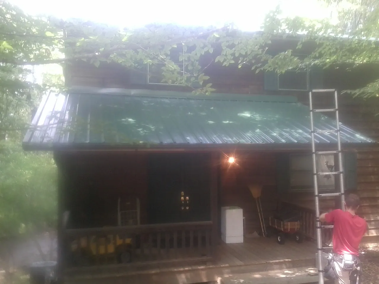 Worker on a ladder at the green porch roof of a wooded cabin in Oriskany
